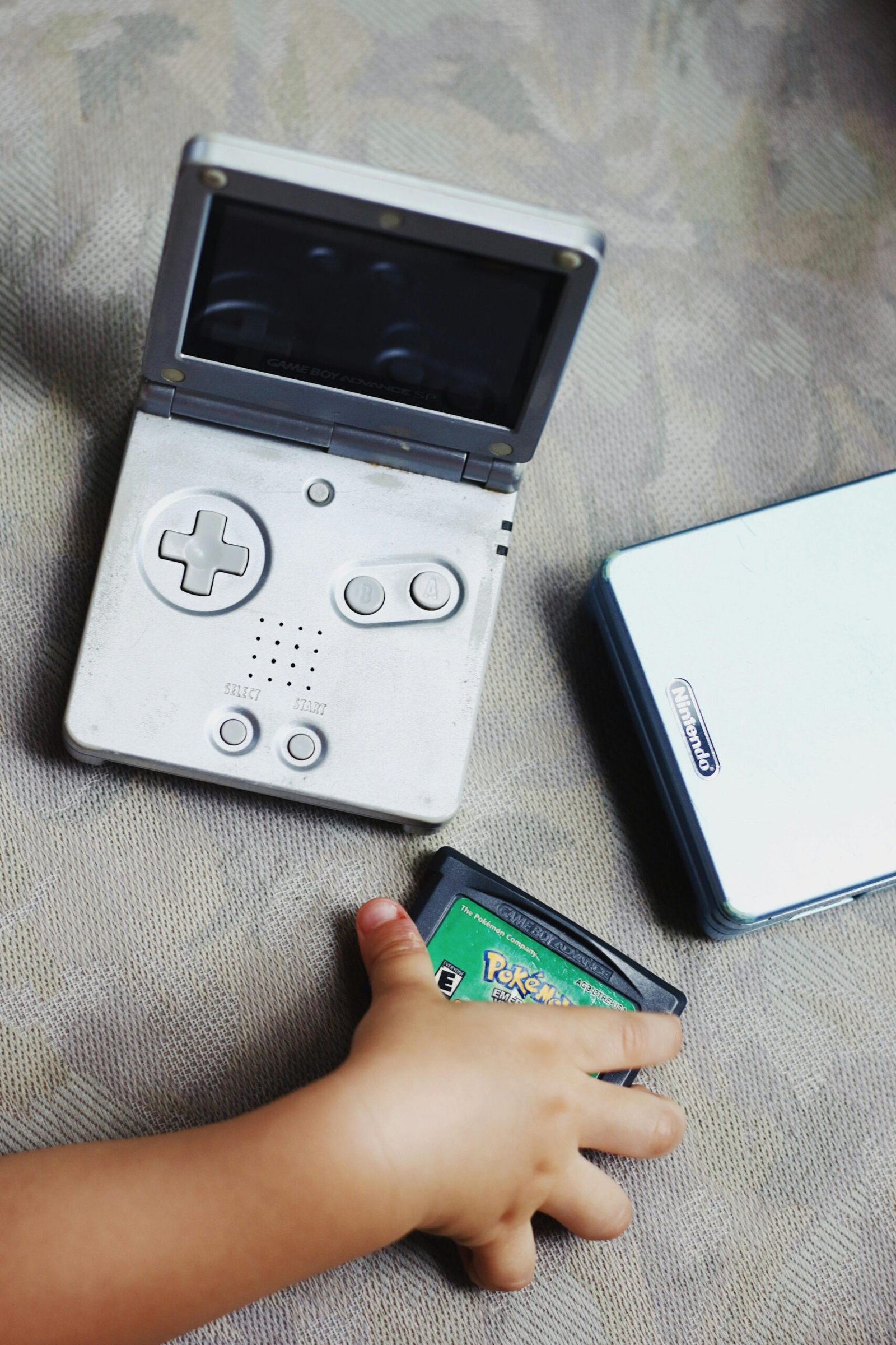 A child's hand holds a vintage Gameboy cartridge next to an open Gameboy SP console.